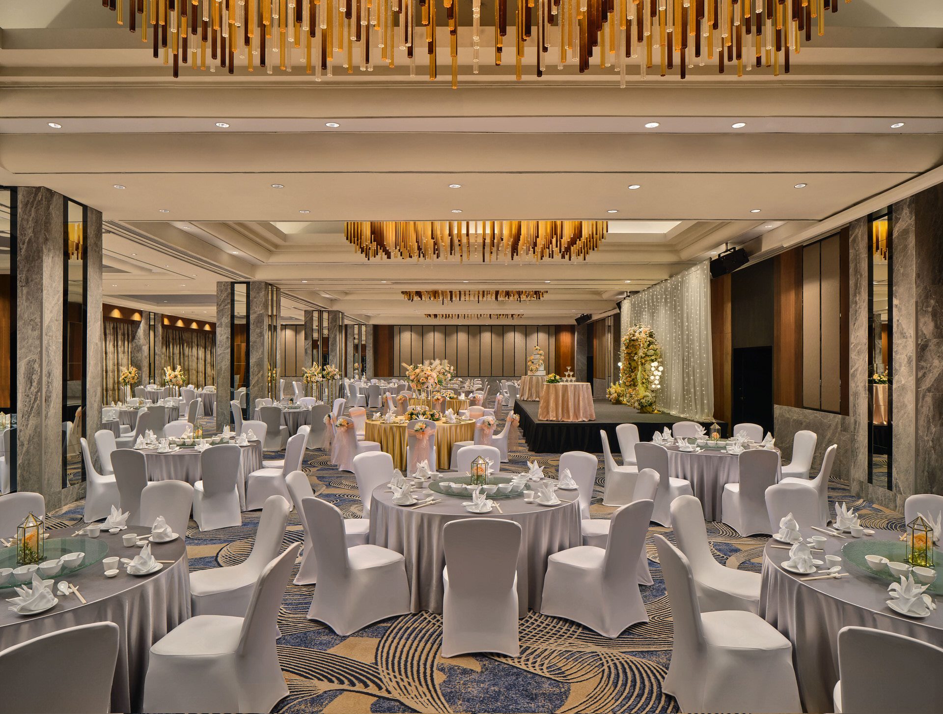 Wide view of a hotel grand ballroom with white banquet rounds, marble columns, patterned carpet, a lit stage backdrop, and gold pendant chandeliers overhead.