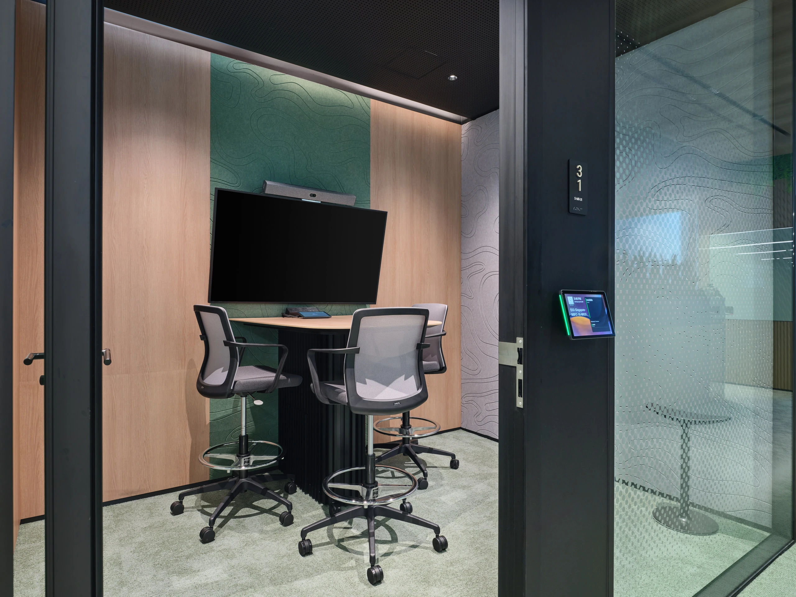 Small glass-walled meeting pod with a wall-mounted monitor, video bar, three grey mesh drafting chairs, teal and wood accent panels, and a digital room sign.
