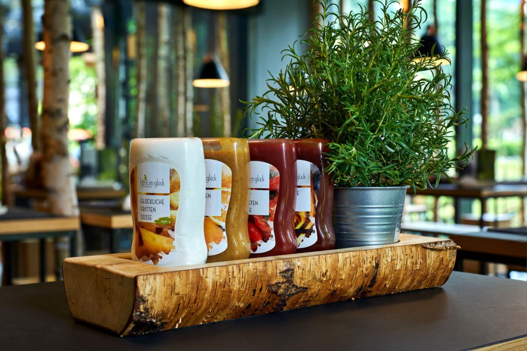 Restaurant photography detail shot of Hans Im Gluck branded merchandise (mugs/bottles) on a wooden surface, part of the commercial shoot.