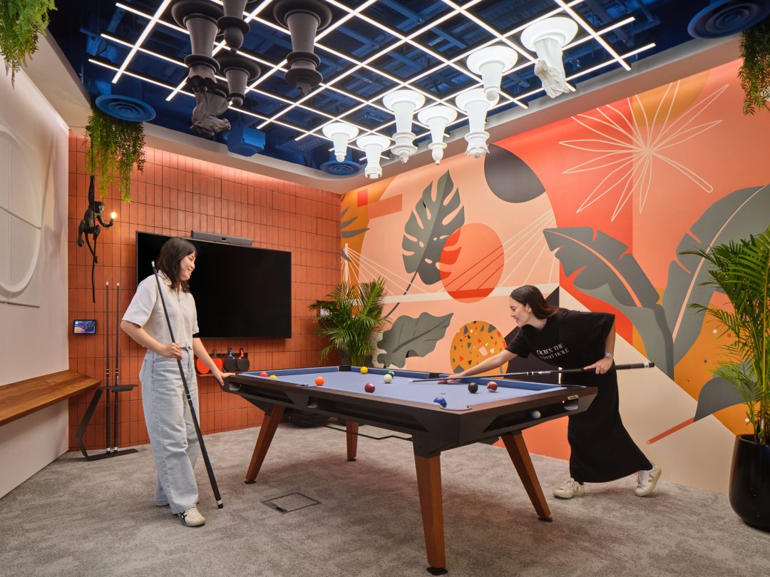 Two employees playing pool in a creative office games room on a convertible conference table, with a unique chess set mounted on the ceiling and a monkey statue in the corner—a prime example of modern workplace photography in Singapore.