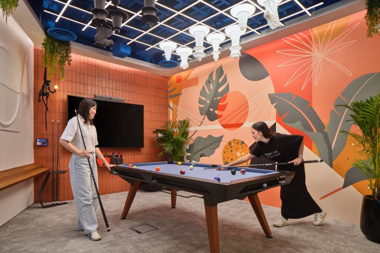 Two employees playing pool in a creative office games room on a convertible conference table, with a unique chess set mounted on the ceiling and a monkey statue in the corner—a prime example of modern workplace photography in Singapore.